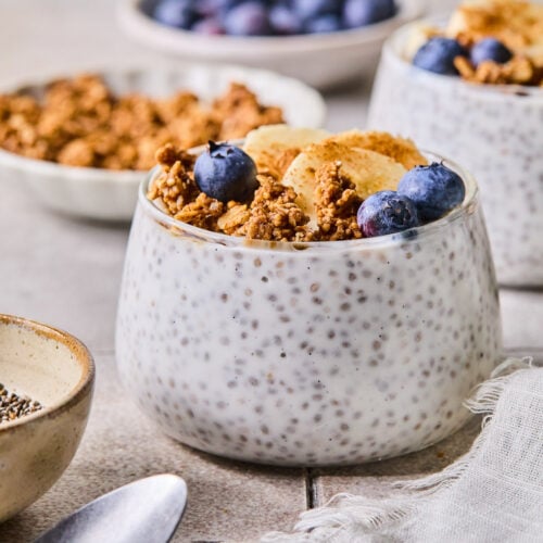 A glass cup with kefir chia pudding topped with granola, blueberries, sliced banana, and cinnamon. Around the cup are a spoon, a bowl of chia seeds, a bowl of granola, a bowl of blueberries, and another cup of kefir pudding.