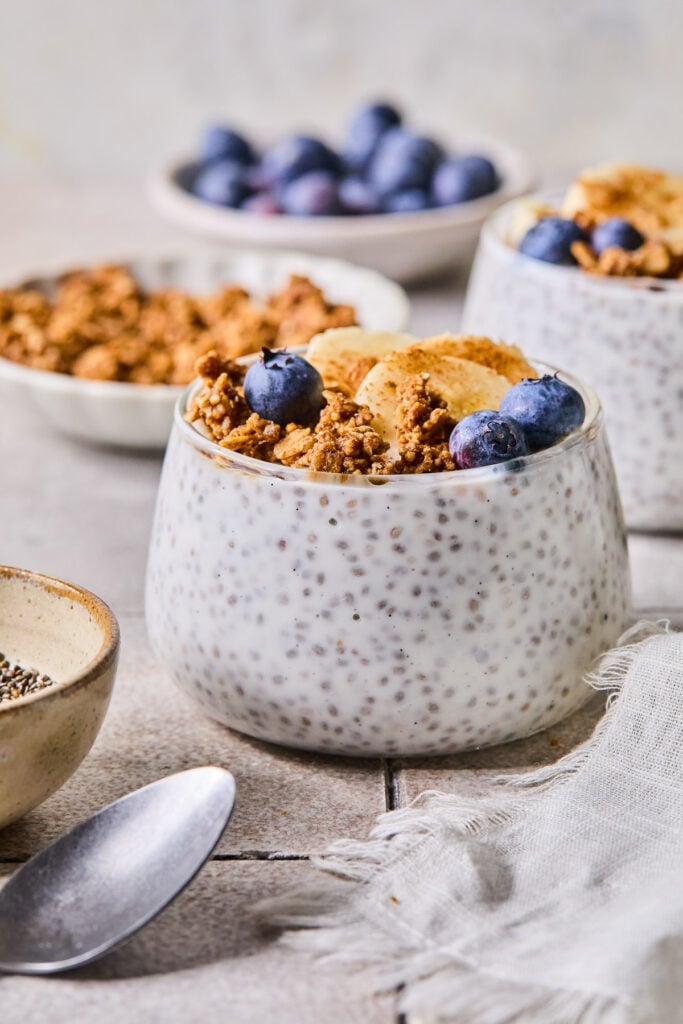 A glass cup with kefir chia pudding topped with granola, blueberries, sliced banana, and cinnamon. Around the cup are a spoon, a bowl of chia seeds, a bowl of granola, a bowl of blueberries, and another cup of kefir pudding.