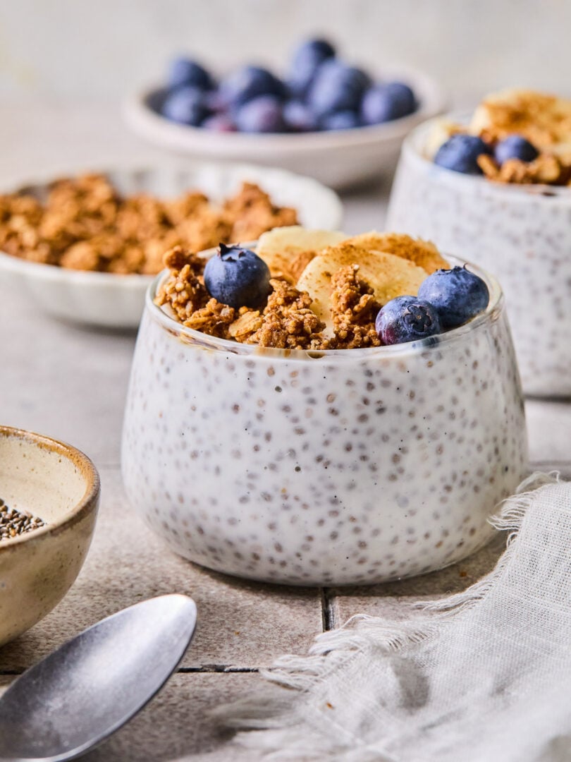 A glass cup with kefir chia pudding topped with granola, blueberries, sliced banana, and cinnamon. Around the cup are a spoon, a bowl of chia seeds, a bowl of granola, a bowl of blueberries, and another cup of kefir pudding.
