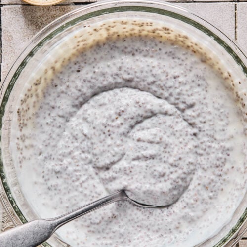 Chia pudding with kefir in a large glass bowl with a spoon in it. Around the bowl is a bowl of chia seeds and a white kitchen towel.