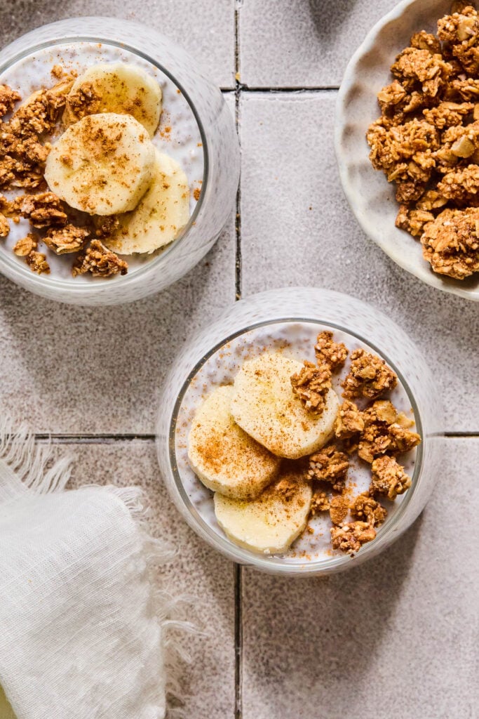 Overhead shot of two glass cups of kefir chia pudding topped with sliced banana, granola, and cinnamon. Around the glasses are a bowl of granola and a white kitchen towel.
