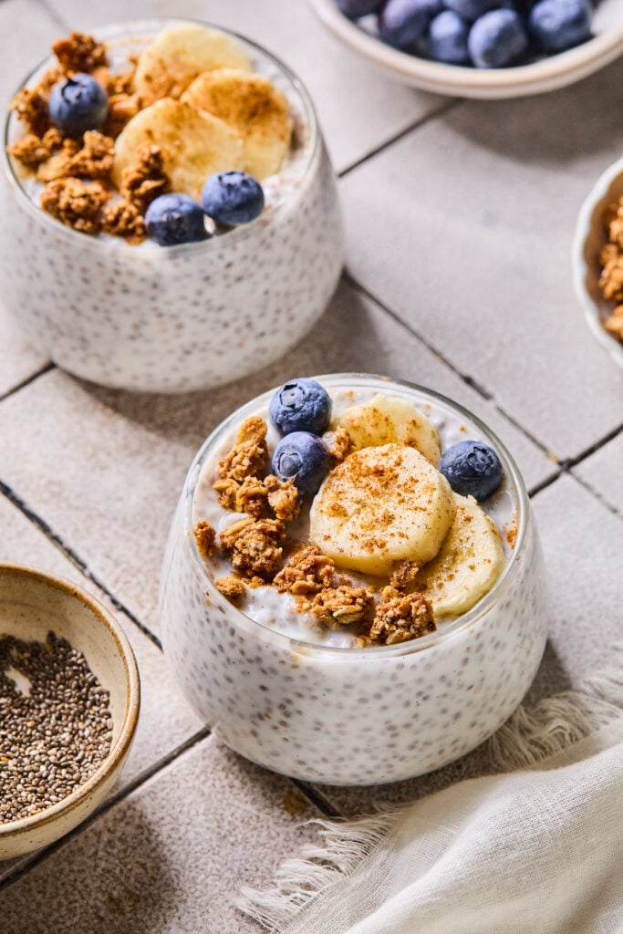 Two glass cups of kefir chia seed pudding topped with granola, sliced banana, blueberries, and cinnamon. Around the bowls are a white kitchen towel, a bowl of chia seeds, a bowl of blueberries, and a bowl of granola.