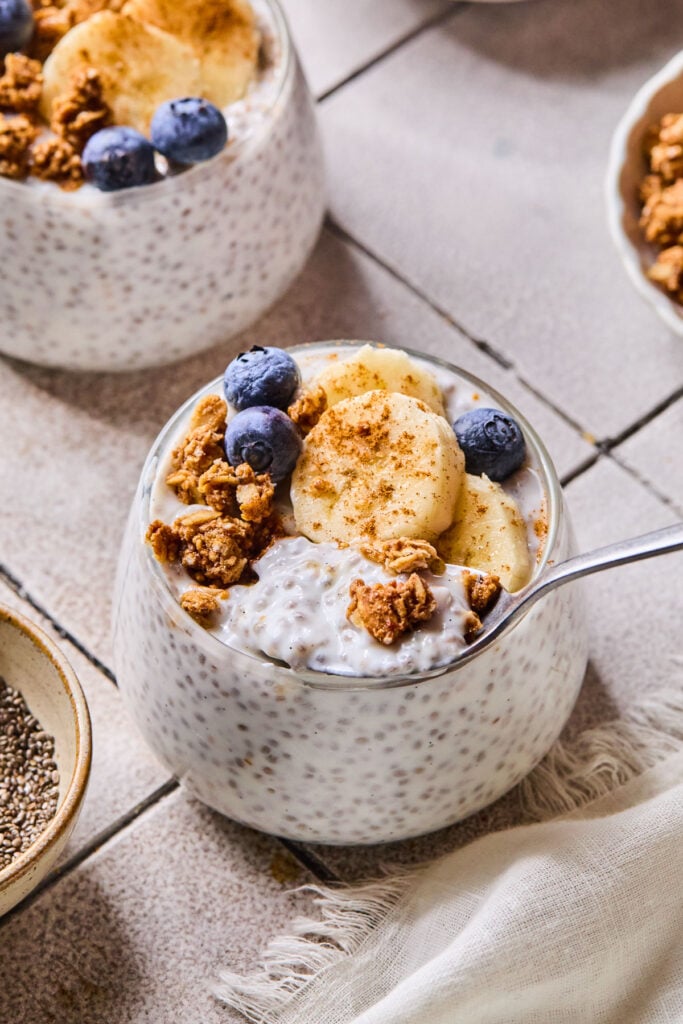 A glass cup of chia kefir pudding topped with granola, blueberries, sliced banana, and cinnamon. A spoon scoops chia pudding from the cup. Around the cup are a white kitchen towel, a bowl of chia seeds, another glass of kefir pudding, and a bowl of granola.