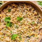 Mexican street corn salad topped with cilantro in a bowl with a spoon in it. Around the bowl are some cilantro leaves and a bowl of chopped cilantro.