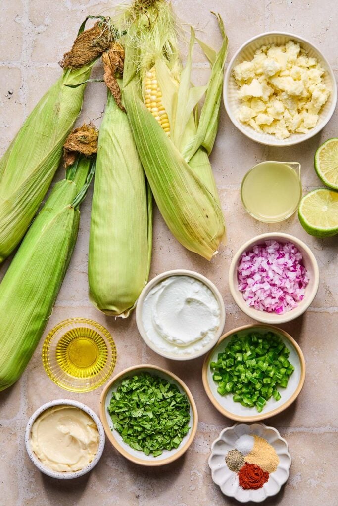 A bowl of cotija cheese, a small cup of lime juice, a lime that has been cut in half, a small bowl of diced red onion, a small bowl of diced jalape&ntilde;o, a small dish with four different piles on spices on it, a small bowl of chopped cilantro, a small bowl of mayo, a small bowl of olive oil, and small bowl of greek yogurt, and 4 ears of corn on a tan tile counter. 