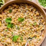 Mexican street corn salad topped with cilantro in a bowl with a spoon in it. Around the bowl are some cilantro leaves, an ear of corn, and a bowl of chopped cilantro.