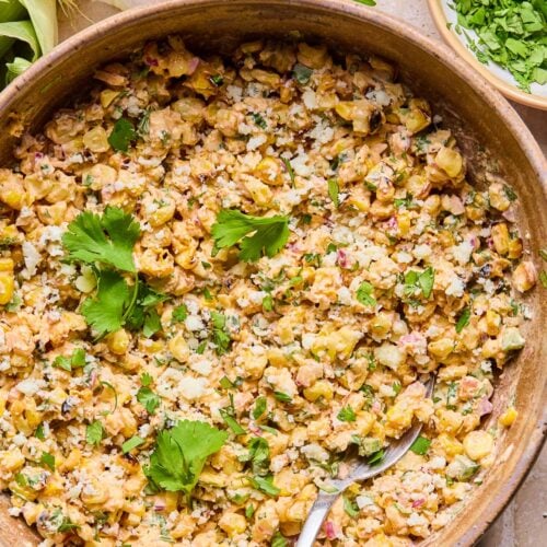 Mexican street corn salad topped with cilantro in a bowl with a spoon in it. Around the bowl are some cilantro leaves, an ear of corn, and a bowl of chopped cilantro.