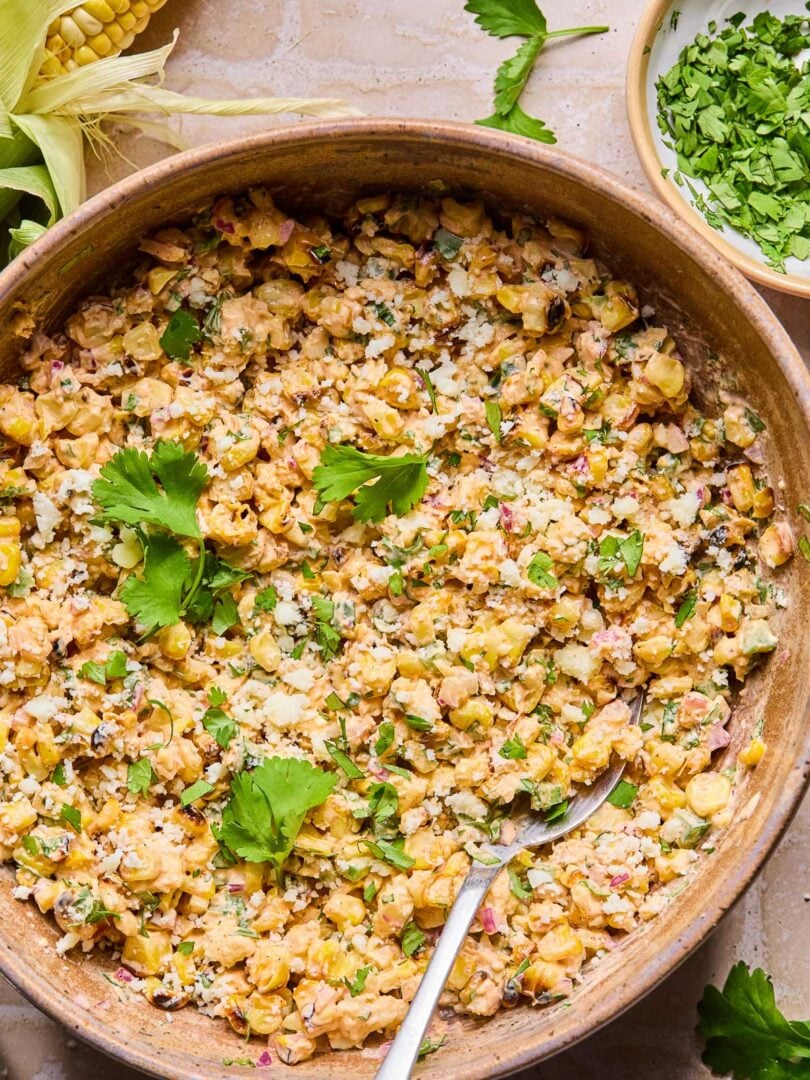 Mexican street corn salad topped with cilantro in a bowl with a spoon in it. Around the bowl are some cilantro leaves, an ear of corn, and a bowl of chopped cilantro.