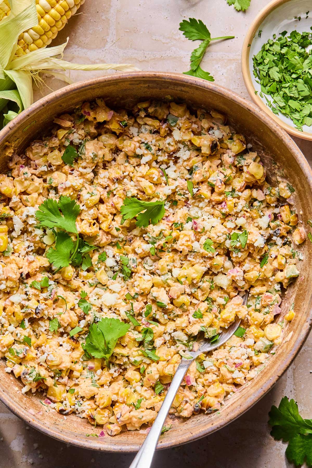 Mexican street corn salad topped with cilantro in a bowl with a spoon in it. Around the bowl are some cilantro leaves, an ear of corn, and a bowl of chopped cilantro.
