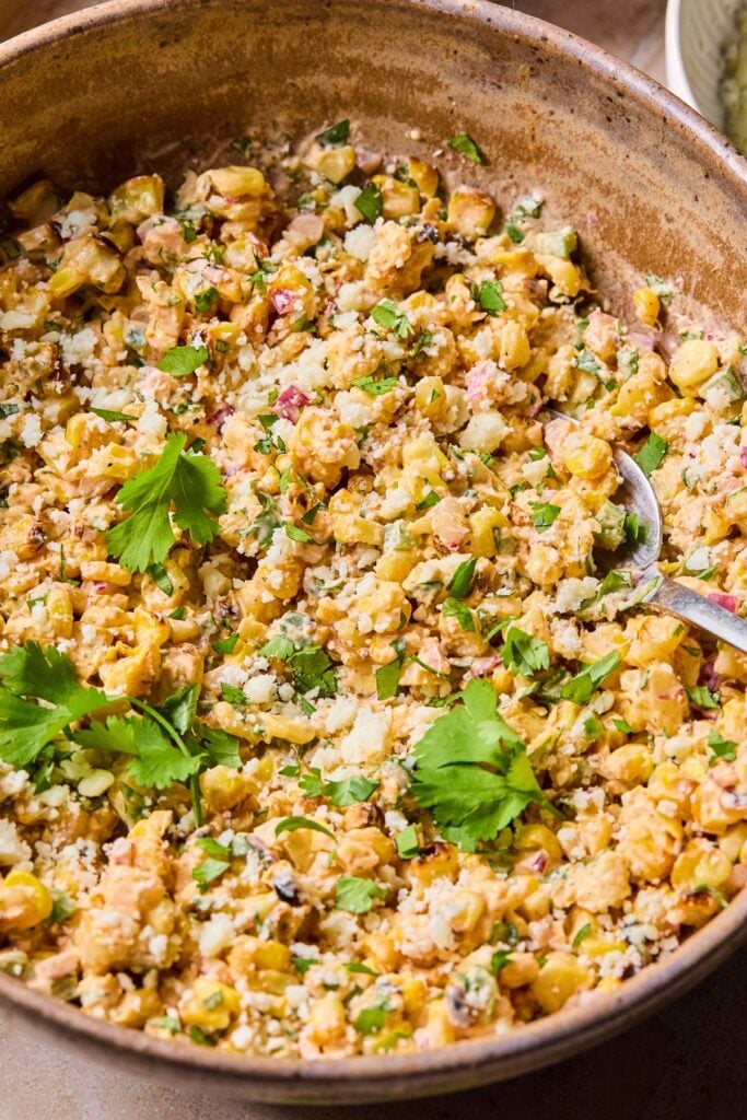 A close-up of a bowl of street corn salad garnished with cilantro, with a spoon in it.