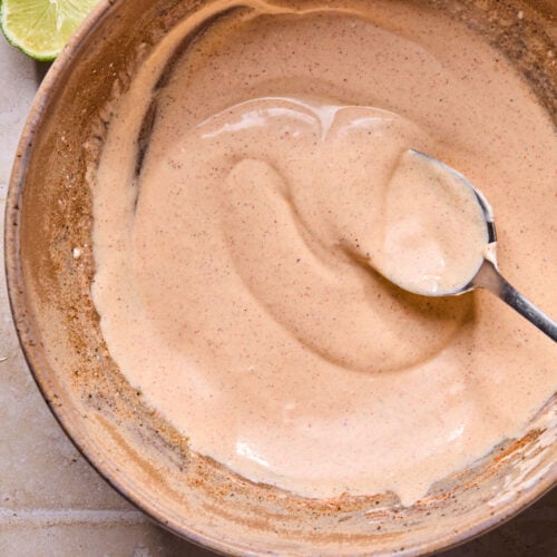 A bowl of a creamy mixture of greek yogurt, mayo, lime juice, and spices with a spoon in it. Next to the bowl is a lime cut in half.