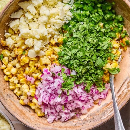 A bowl of creamy dressing topped with piles of charred corn kernels, cotija cheese, diced jalape&ntilde;o, chopped cilantro, and diced red onion before it is mixed. A spoon is in the bowl as well. Around the bowl is a bowl of cotija cheese, a lime cut in half, and a bowl of cilantro.