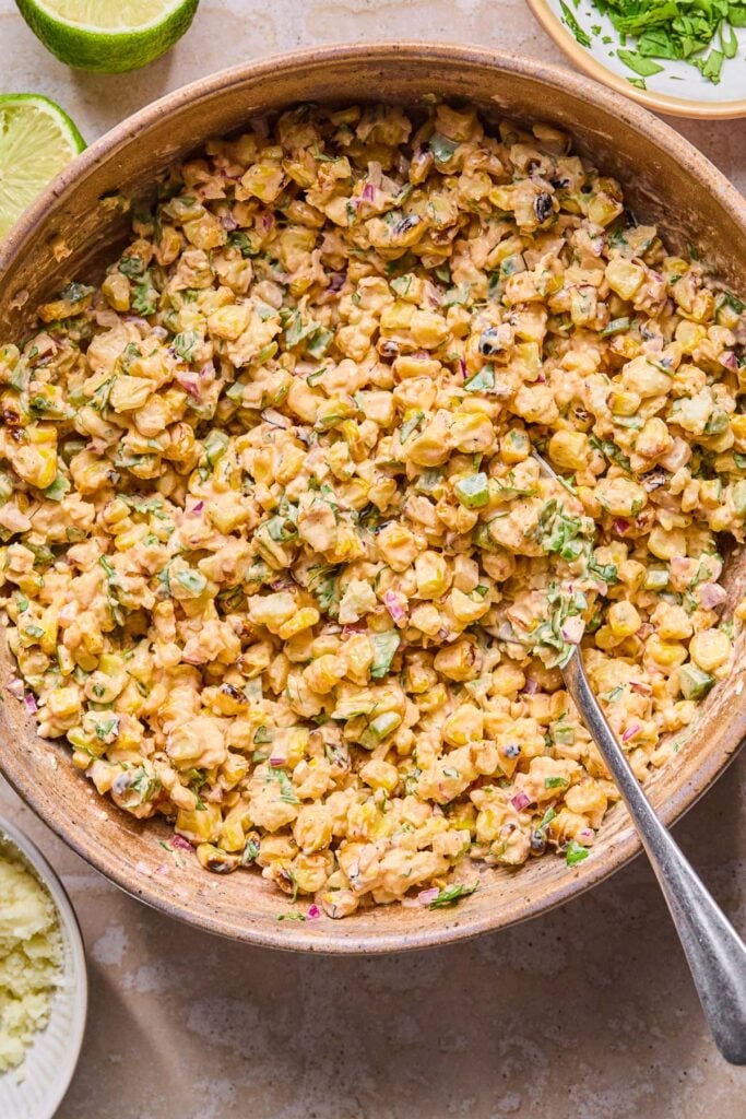Mexican street corn salad in a bowl with a spoon in it. Around the bowl is a bowl of cotija cheese, a lime cut in half, and a bowl of cilantro.