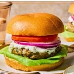 Greek turkey burger with lettuce, tomato, red onion, and tzatziki sauce on a bun on a plate. Around the plate are a bowl of tzatziki sauce, a striped towel, a glass of water, and another burger on a plate.