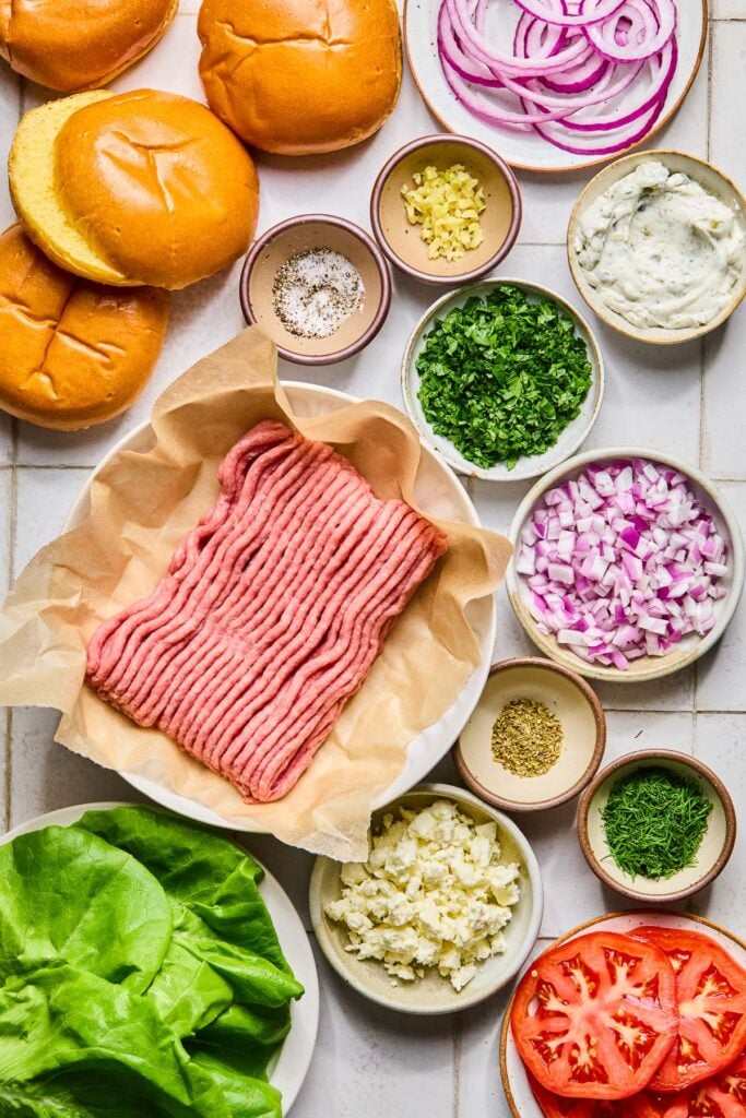 A plate of sliced red onion, a small bowl of minced garlic, a bowl of tzatziki sauce, of bowl of chopped parsley, a bowl of diced red onion, a small bowl of dried oregano, a small bowl of chopped dill, a plate of sliced tomatoes, a bowl of crumbled feta, a plate of lettuce leaves, a plate of a pound of raw ground turkey, a small bowl of salt and pepper, and some hamburger buns on a grey tile counter.