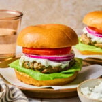 Greek turkey burger with lettuce, tomato, red onion, and tzatziki sauce on a bun on a plate. Around the plate are a bowl of tzatziki sauce, a striped towel, a glass of water, and another burger on a plate.