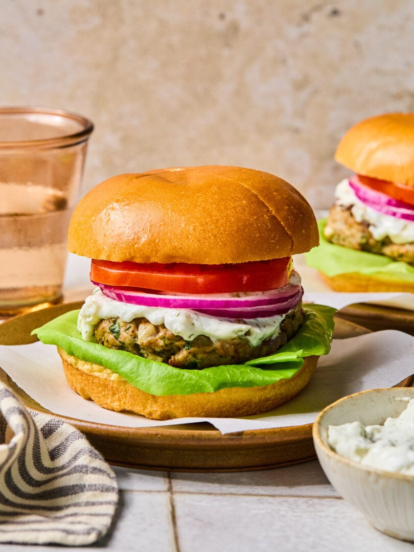 Greek turkey burger with lettuce, tomato, red onion, and tzatziki sauce on a bun on a plate. Around the plate are a bowl of tzatziki sauce, a striped towel, a glass of water, and another burger on a plate.