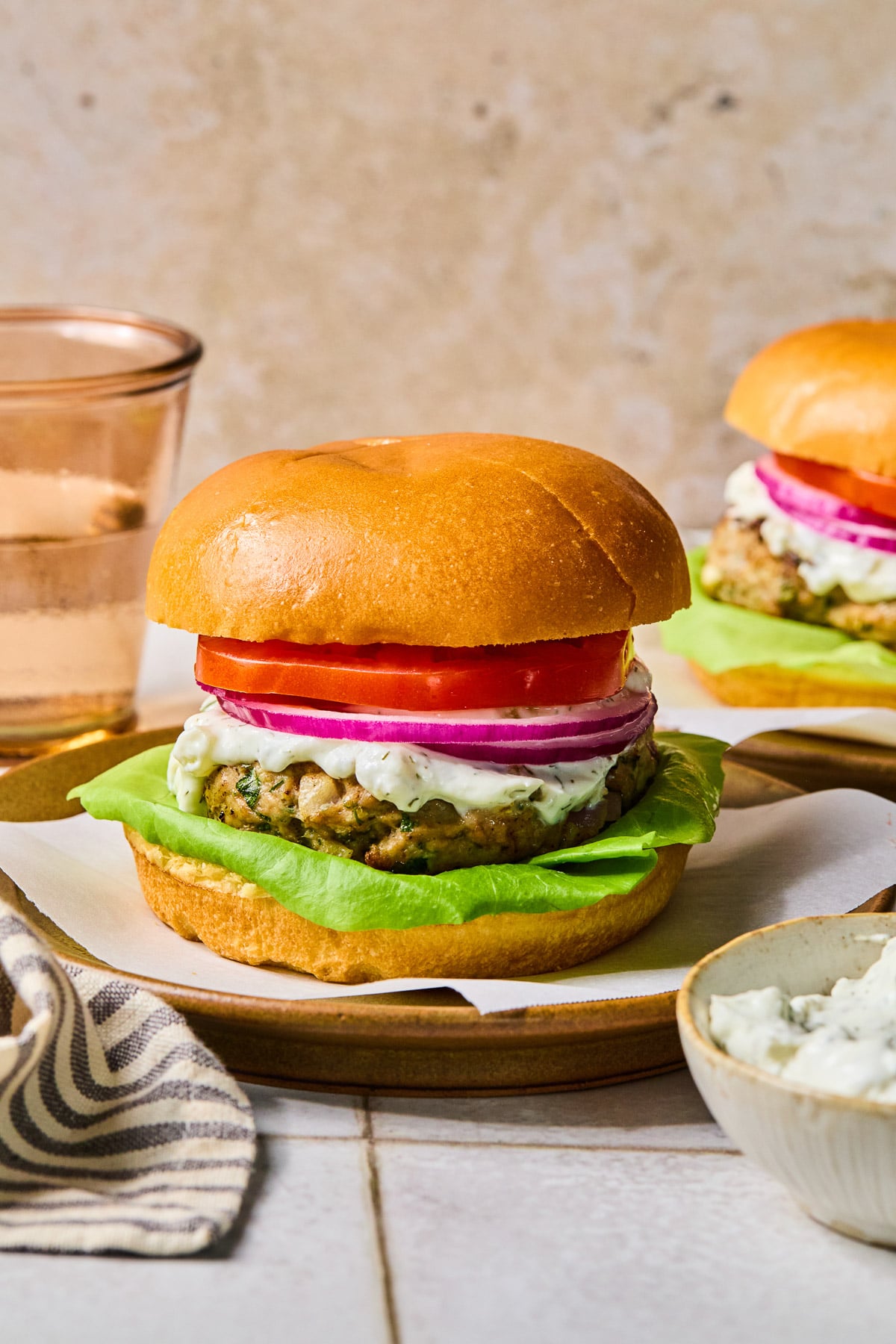 Greek turkey burger with lettuce, tomato, red onion, and tzatziki sauce on a bun on a plate. Around the plate are a bowl of tzatziki sauce, a striped towel, a glass of water, and another burger on a plate.