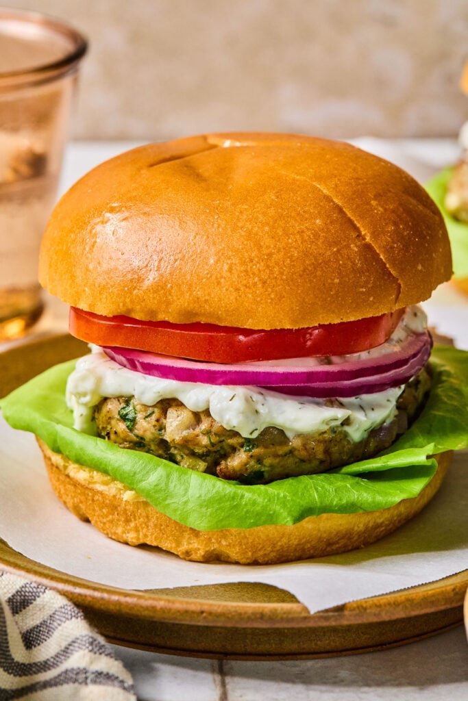 Close-up of a plate with greek turkey burger on a bun with lettuce, tomato, red onion, and tzatziki sauce. In front of the plate is a striped towel, and behind the plate is a glass of water.