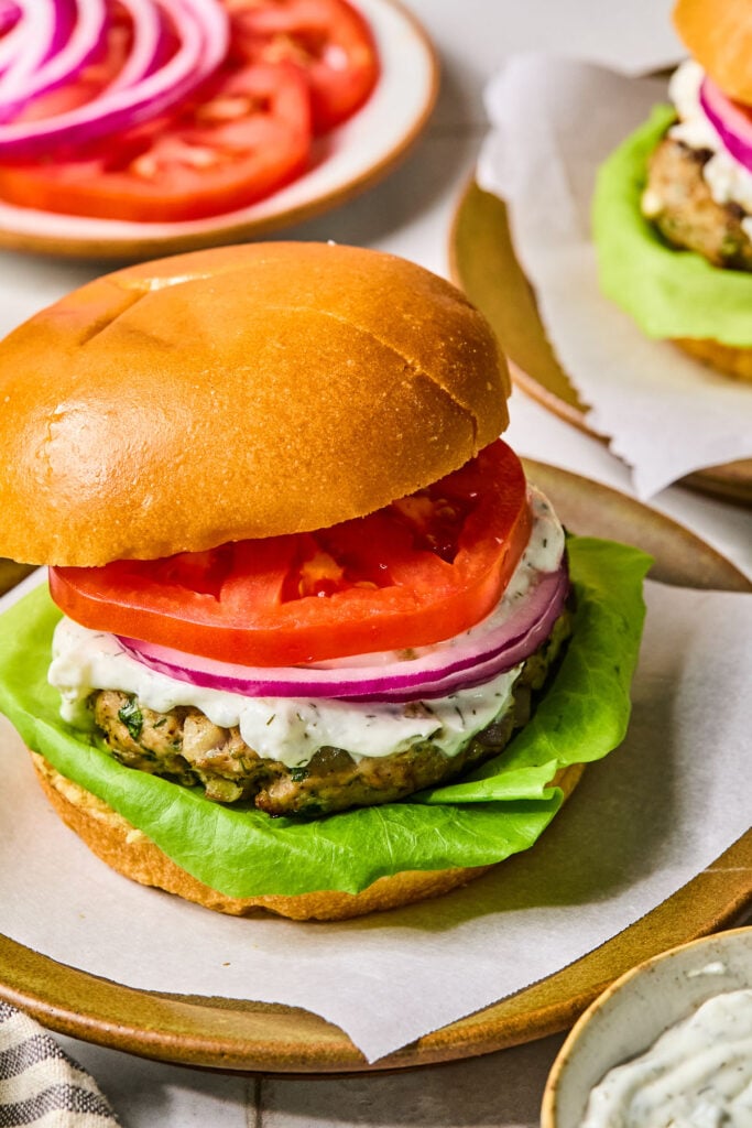 A plate of a turkey feta burger on a bun with lettuce, tomato, red onion, and tzatziki sauce. Around the plate is a bowl of tzatziki sauce, a striped towel, a plate of sliced tomato and red onion, and a plate with another burger on it.