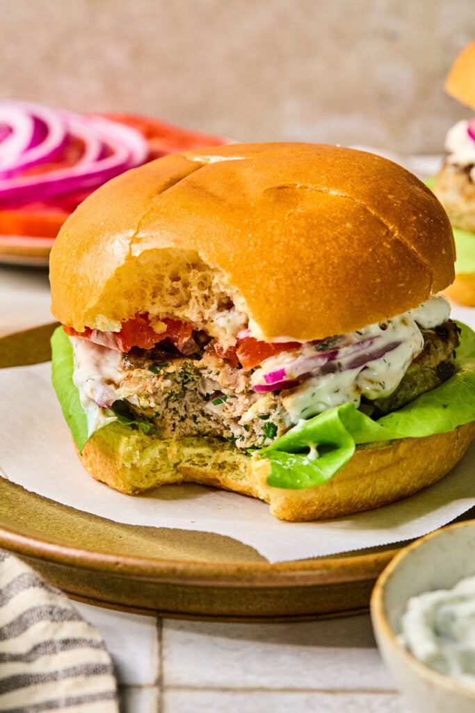 Greek turkey burger with lettuce, tomato, red onion, and tzatziki sauce in a bun with a bite taken out of it. Around the plate is a bowl of tzatziki sauce, a striped towel, and a plate with sliced tomato and red onion.