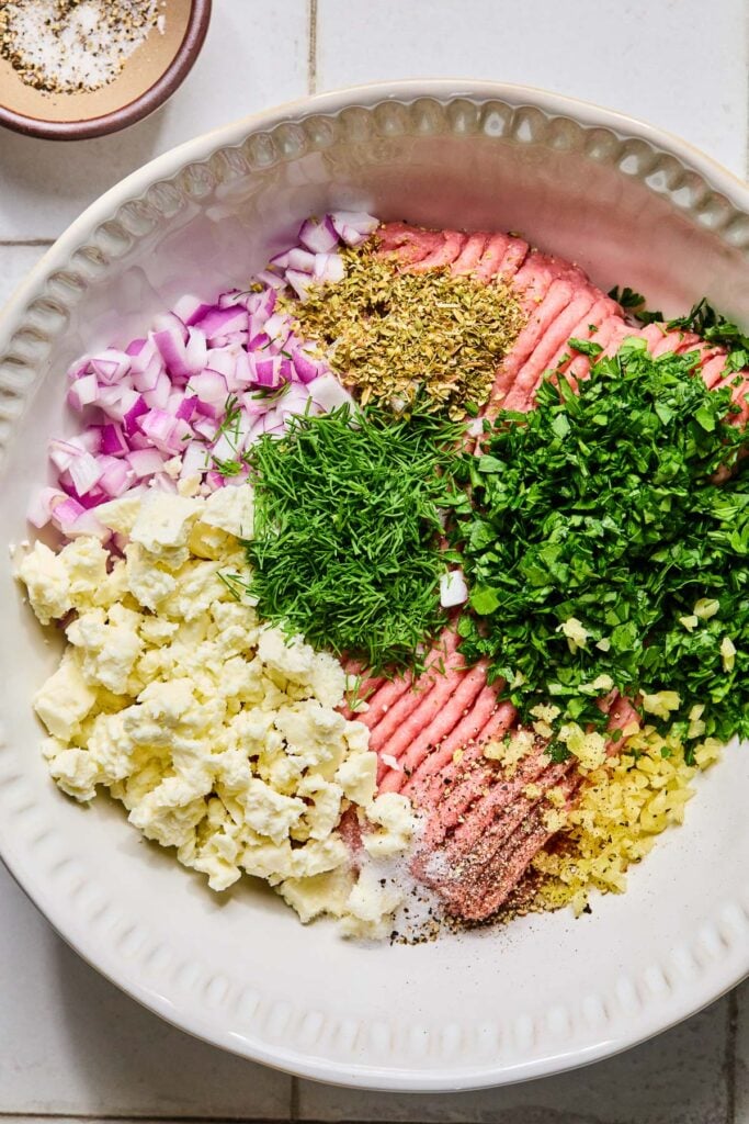 A large bowl of raw ground turkey, crumbled feta cheese, diced red onion, chopped fresh herbs, minced garlic, spices, salt, and pepper before it is mixed. Next to the bowl is a small bowl of salt and pepper.