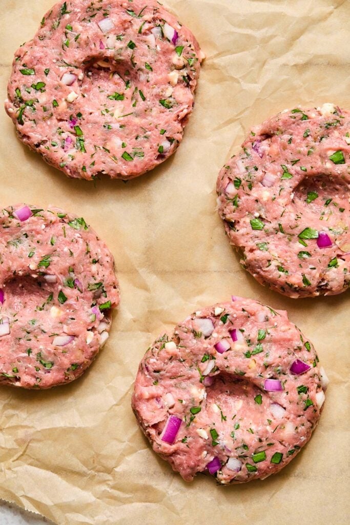 Raw turkey burger patties with onion, feta, and fresh herbs on a piece of parchment paper.