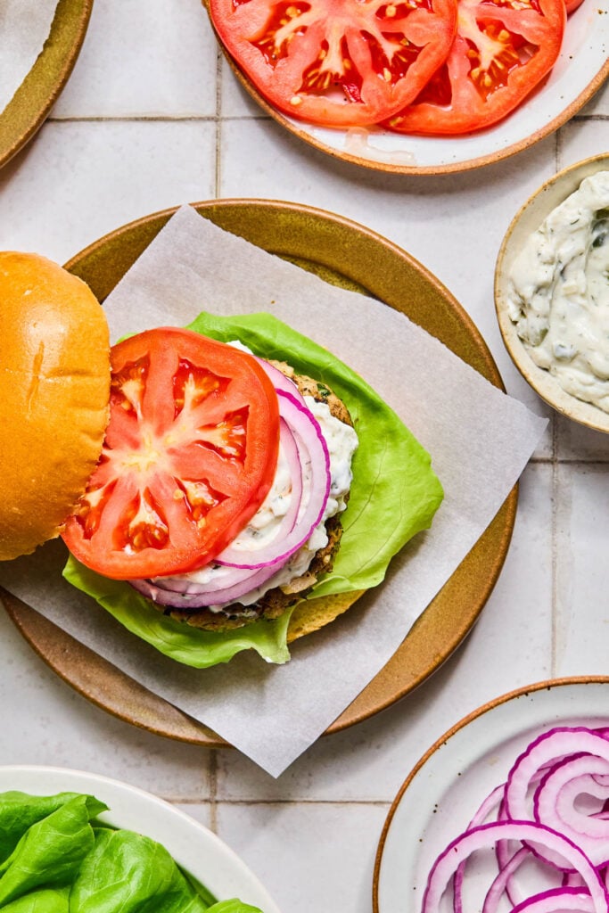 Overhead shot of a plate with a burger being assembled on it. It has a bun, lettuce, a burger patty, tzatziki sauce, red onion, tomato, and the top of the bun. Around the plate are a plate of sliced tomato, a bowl of tzatziki sauce, a plate of sliced red onion, and a plate of lettuce.