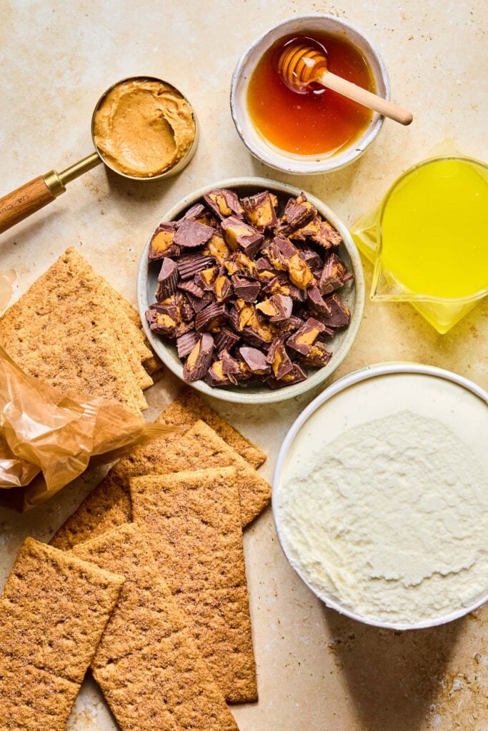 A small bowl of honey with a honey dipper in it, a measuring cup of melted butter, a large pint of vanilla ice cream, graham crackers coming out of a wrapper, a bowl of chopped peanut butter cups, and a 1/2 cup of peanut butter on a tan concrete counter.