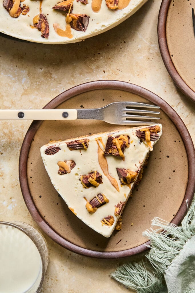 A slice of Reese’s peanut butter ice cream cake on a plate with a fork on it. Around the plate is a green kitchen towel, a glass of milk, the rest of the cake, and another small plate.