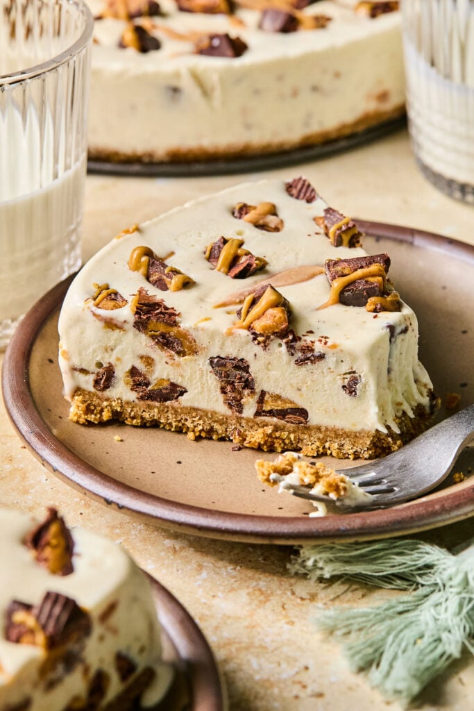 A slice of Reese’s peanut butter cup ice cream cake on a plate. A bite is taken out of the cake, and a fork is resting on the plate. Around the plate are a green kitchen towel, a plate with another piece of cake on it, two glasses of milk, and the rest of the cake.