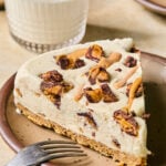 A slice of Reese's ice cream cake on a plate with a fork resting on the plate. Behind the plate is a glass of milk and another plate with a piece of cake.