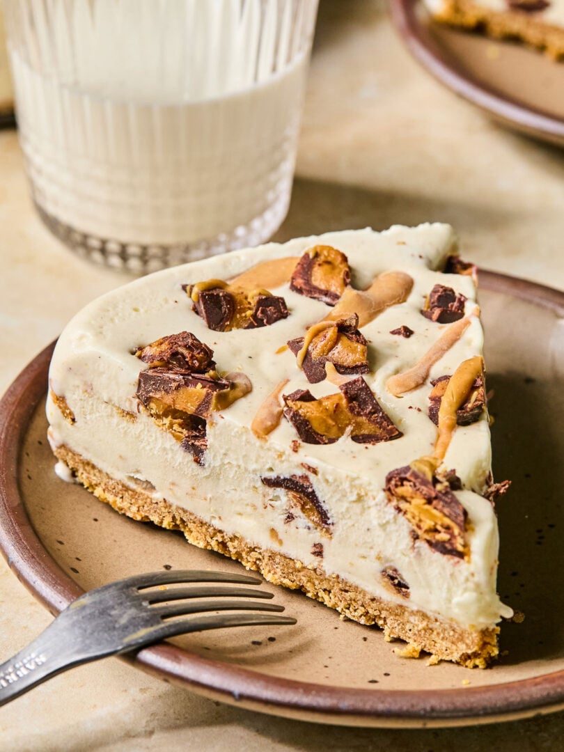 A slice of Reese's ice cream cake on a plate with a fork resting on the plate. Behind the plate is a glass of milk and another plate with a piece of cake.