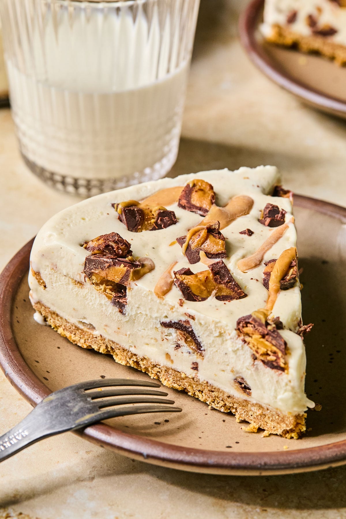 A slice of Reese's ice cream cake on a plate with a fork resting on the plate. Behind the plate is a glass of milk and another plate with a piece of cake.
