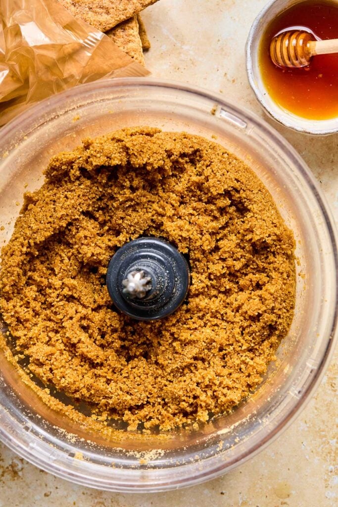 A food processor filled with a mixture of crushed graham crackers, honey, and butter. Next to the food processor are some graham crackers and a bowl of honey with a honey dipper in it.