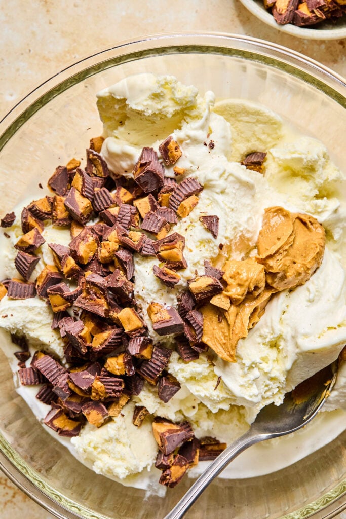 A bowl of vanilla ice cream topped with chopped peanut butter cups and peanut butter before mixing. A spoon is also in the bowl. Next to the bowl is a small bowl with chopped peanut butter cups.