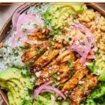 Street corn chicken rice bowl with romaine, sliced avocado, and pickled onions. It is garnished with cilantro and cotija cheese. Around the bowl is a yellow kitchen towel, a bowl of chopped cilantro, and a bowl of chopped romaine.