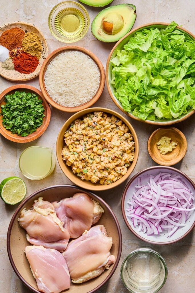 An avocado that has been cut in half, a bowl of chopped romaine, a bowl of minced garlic, a bowl of sliced red onion, a jar of white vinegar, a plate of raw chicken thighs, half a lime, a small jar of lime juice, a bowl of Mexican street corn salad, a bowl of chopped cilantro, a bowl of uncooked white rice, a plate of piles of different spices, and a small bowl of olive oil on a tan tile counter.