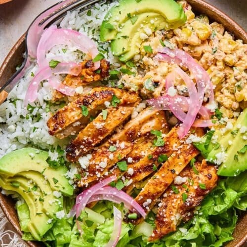 Street corn chicken rice bowl with romaine, sliced avocado, and pickled onions. It is garnished with cilantro and cotija cheese. Around the bowl is a yellow kitchen towel, a bowl of chopped cilantro, and a bowl of chopped romaine.