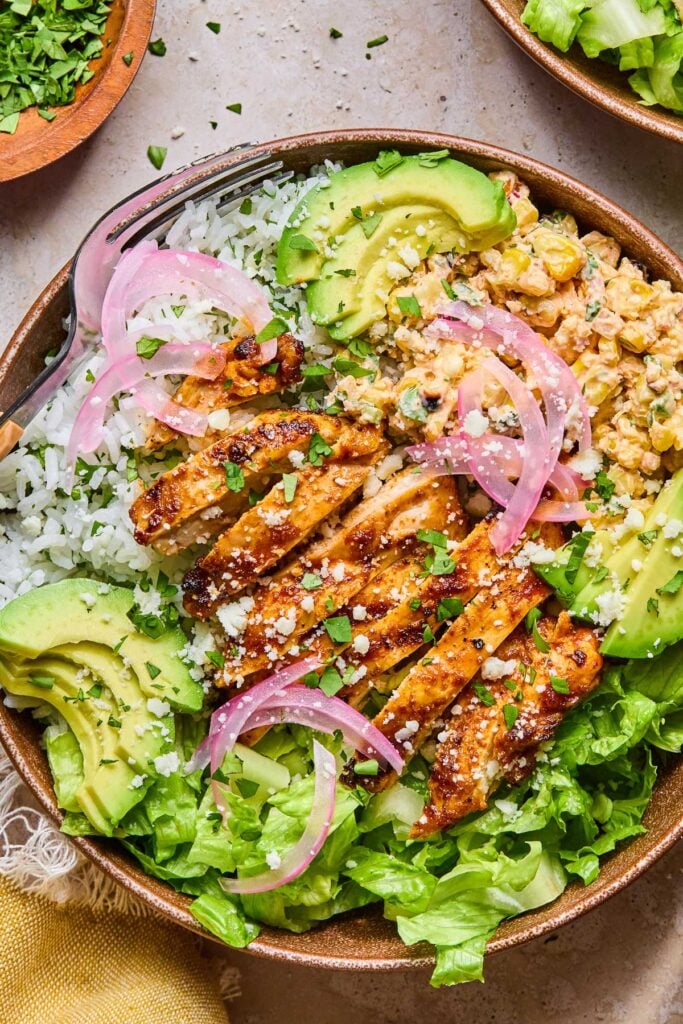 Street corn chicken rice bowl with romaine, sliced avocado, and pickled onions. It is garnished with cilantro and cotija cheese. Around the bowl is a yellow kitchen towel, a bowl of chopped cilantro, and a bowl of chopped romaine.