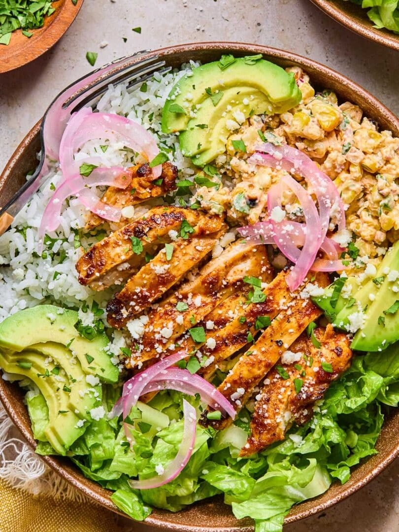 Street corn chicken rice bowl with romaine, sliced avocado, and pickled onions. It is garnished with cilantro and cotija cheese. Around the bowl is a yellow kitchen towel, a bowl of chopped cilantro, and a bowl of chopped romaine.