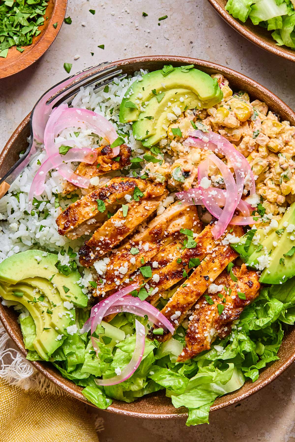 Street corn chicken rice bowl with romaine, sliced avocado, and pickled onions. It is garnished with cilantro and cotija cheese. Around the bowl is a yellow kitchen towel, a bowl of chopped cilantro, and a bowl of chopped romaine.