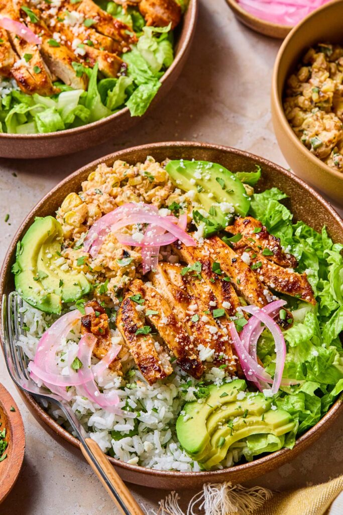 Mexican street corn chicken bowl with cilantro lime rice, grilled chicken, street corn salad, romaine, avocado, and pickled onions. Af ork is in the bowl too. Around the bowl is another street corn chicken bowl, a small dish of pickled onions, a bowl of Mexican street corn, and a yellow kitchen towel.