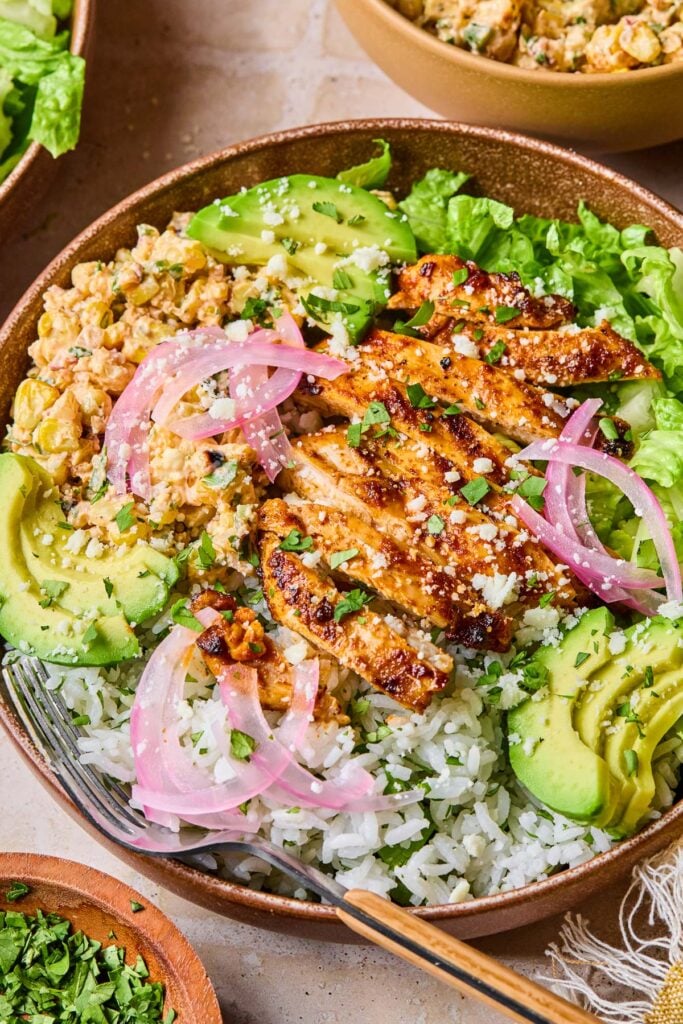 Street corn chicken bowl with cilantro lime rice, grilled chicken, Mexican street corn salad, romaine, avocado, and pickled onions. It is garnished with cilantro and cotija cheese. Around the bowl are a bowl of chopped cilantro, a bowl of chopped romaine, a bowl of street corn salad, and a kitchen towel.