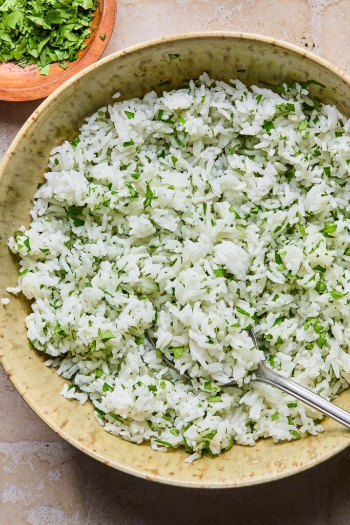 A bowl filled with cilantro lime rice with a fork in it. Next to the bowl is a bowl of chopped cilantro.