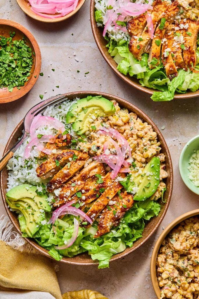 Two street corn chicken bowls with grilled chicken, cilantro lime rice, Mexican street corn salad, romaine, avocado, and pickled onions. Around the bowls are a small bowl of cotija cheese, a bowl of street corn salad, a yellow kitchen towel, a bowl of chopped cilantro, and a bowl of pickled onions.