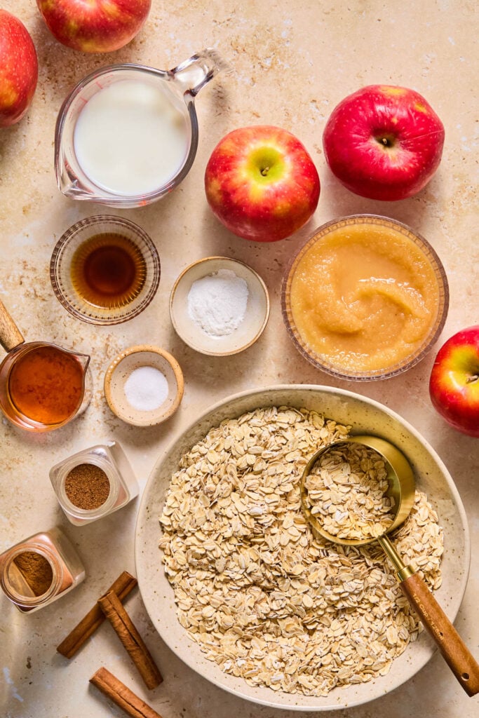 A bowl of applesauce, a large bowl of rolled oats with a measuring cup in it, some cinnamon sticks, two opened spice jars, a small bowl of salt, a measuring cup of maple syrup, a small bowl of baking powder, a small bowl of vanilla extract, a measuring cup of milk, and a handful of fresh apples on a tan concrete counter.