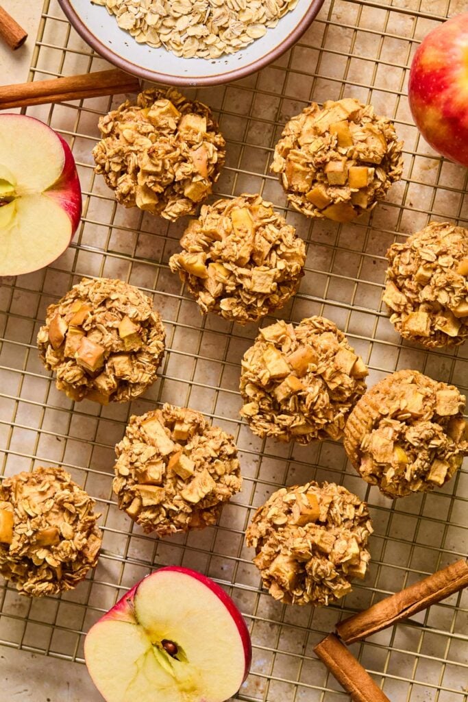 A wire cooling rack with apple cinnamon oatmeal muffins spread across it. Also on the rack are an apple cut in half, some cinnamon sticks, a bowl of rolled oats, and a whole apple.