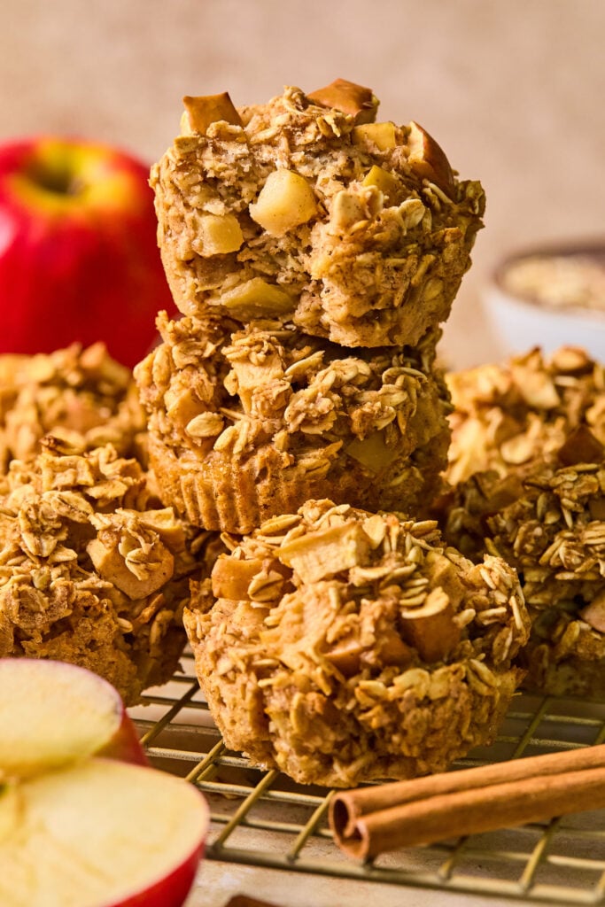 A wire cooling rack with healthy apple muffins on it, with three of them stacked on top of each other. The top one has a bite taken out of it. Around the muffins are a cinnamon stick, an apple cut in half, a whole apple, and a bowl of rolled oats.