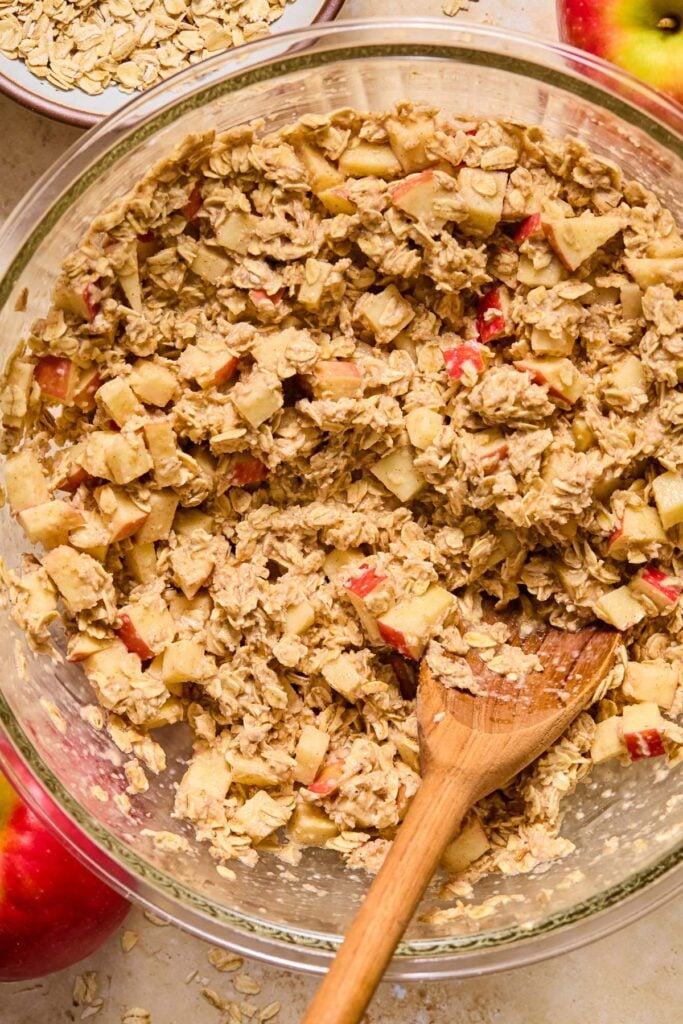 Glass mixing bowl with a wet batter of oats and diced apples with a wooden spoon in it. Around the bowl are two apples and a bowl of rolled oats.